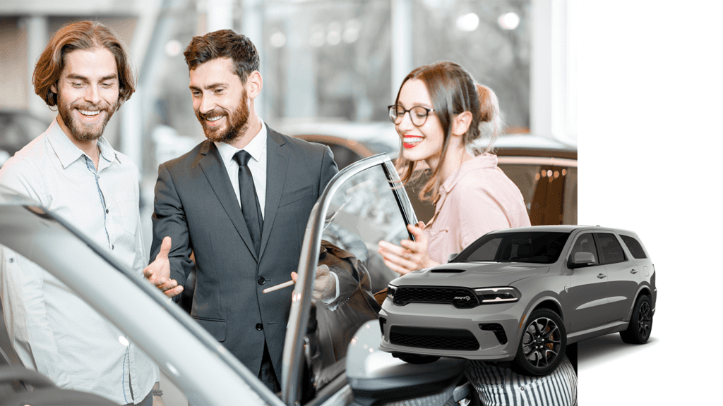 Best Time To Buy At Carolina CDJR in Elizabeth City, NC | 2022 Dodge Durango in the color grey with a background photo of a sales person showing a man and woman some new car options