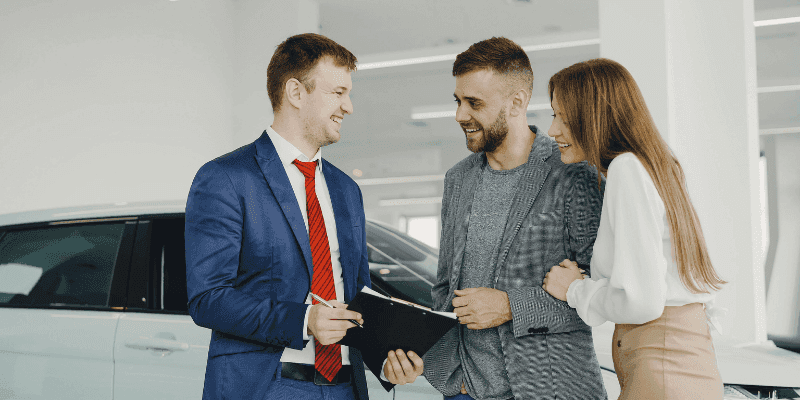 Car salesperson reviewing paperwork with a smiling couple at a dealership