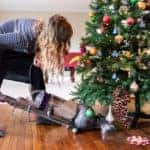 Young woman cleaning with vacuum cleaner, vacuuming under Christmas Tree needles with New Years ornaments on hardwood wooden floor