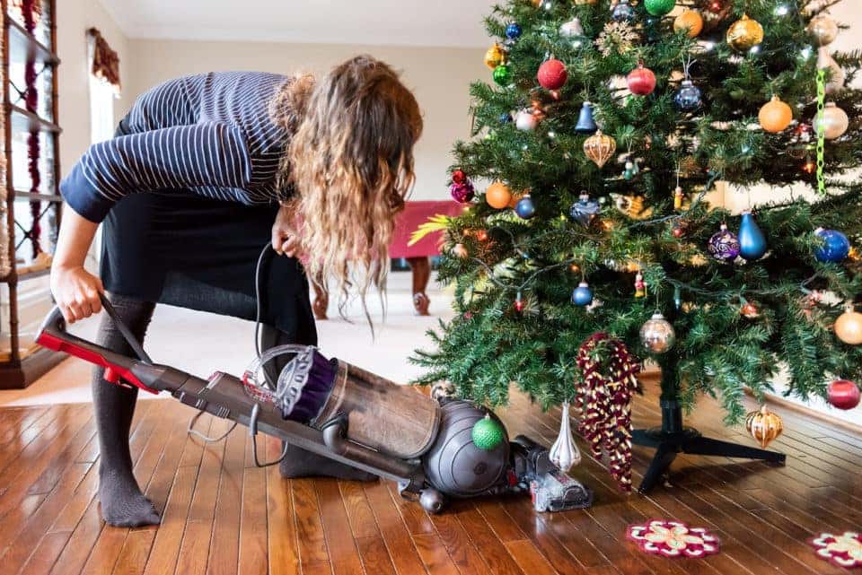 Young woman cleaning with vacuum cleaner, vacuuming under Christmas Tree needles with New Years ornaments on hardwood wooden floor