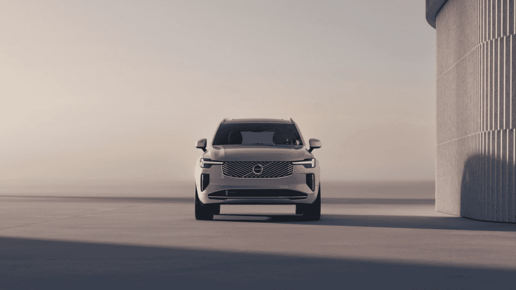 Frontal view of the 2025 Volvo XC90 parked near a modern building in soft sunset lighting, showcasing its sleek grille design.