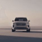 Frontal view of the 2025 Volvo XC90 parked near a modern building in soft sunset lighting, showcasing its sleek grille design.