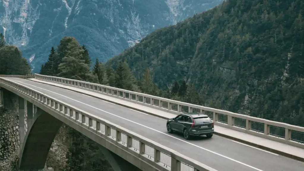 Rear view of the 2025 Volvo XC60 driving across a mountain bridge, emphasizing engine options and strong performance output.