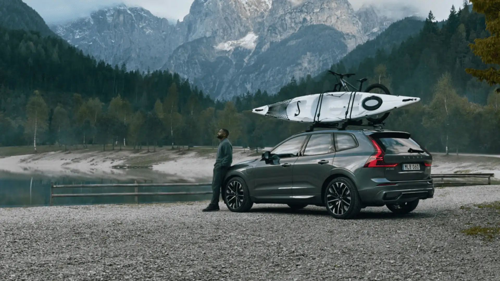 2025 Volvo XC60 SUV parked by a scenic mountain lake in Bristol, TN, equipped with a roof rack carrying a kayak and bike, highlighting its versatility and adventure-ready design.