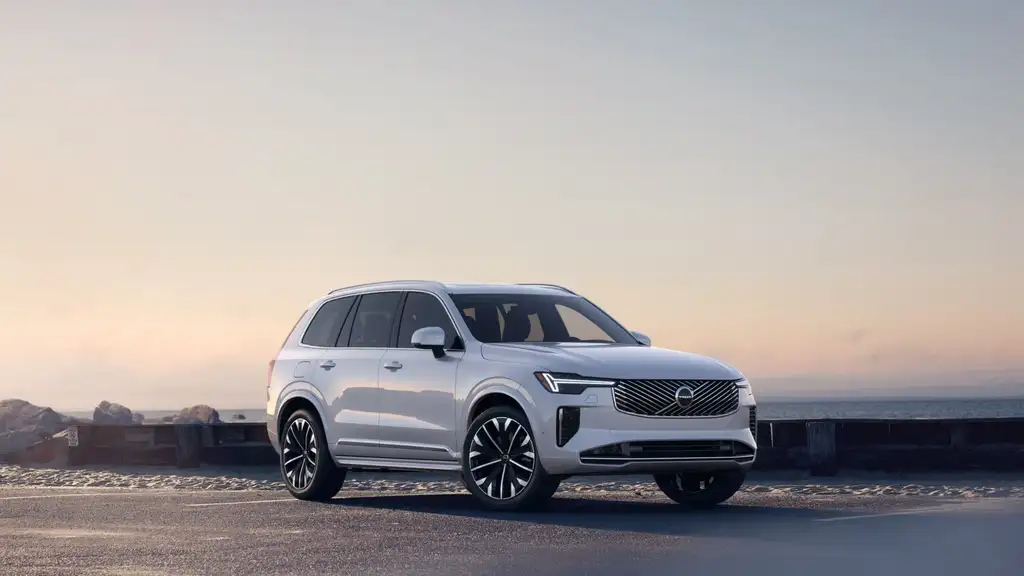 2026 Volvo XC90 Plug-In Hybrid in white with large alloy wheels parked along a coastal road at sunset, compared to Toyota Highlander Hybrid for Johnson City TN drivers