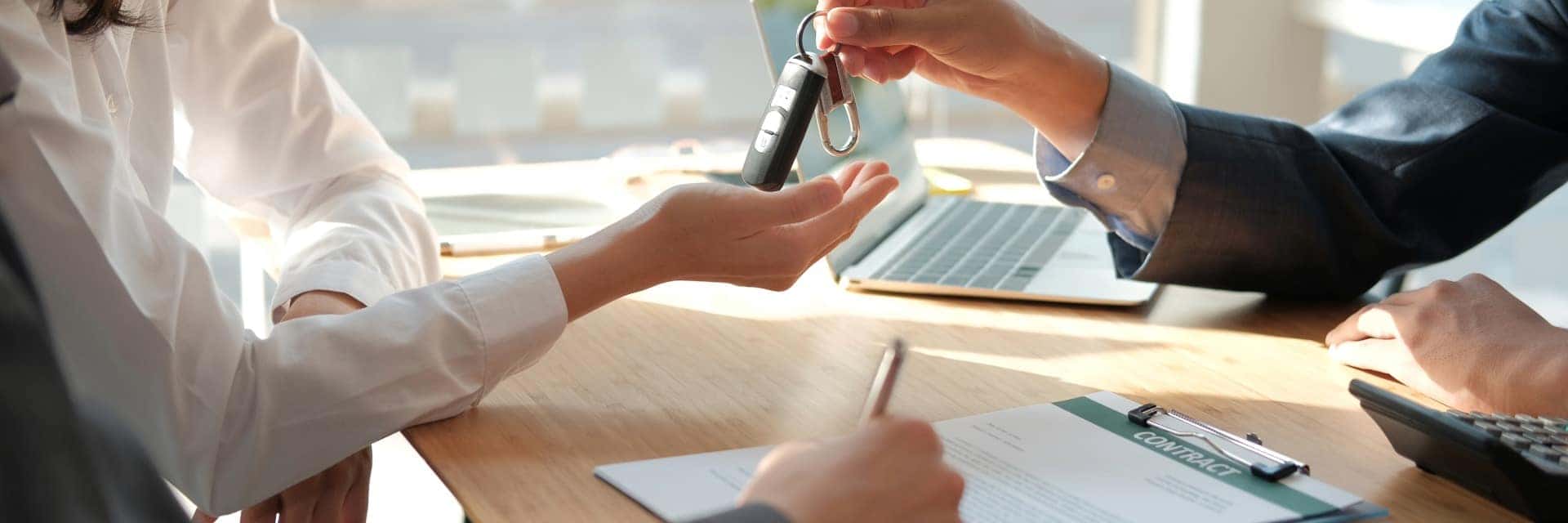Person handing a car key across a desk to a customer, with paperwork and a laptop on the desk