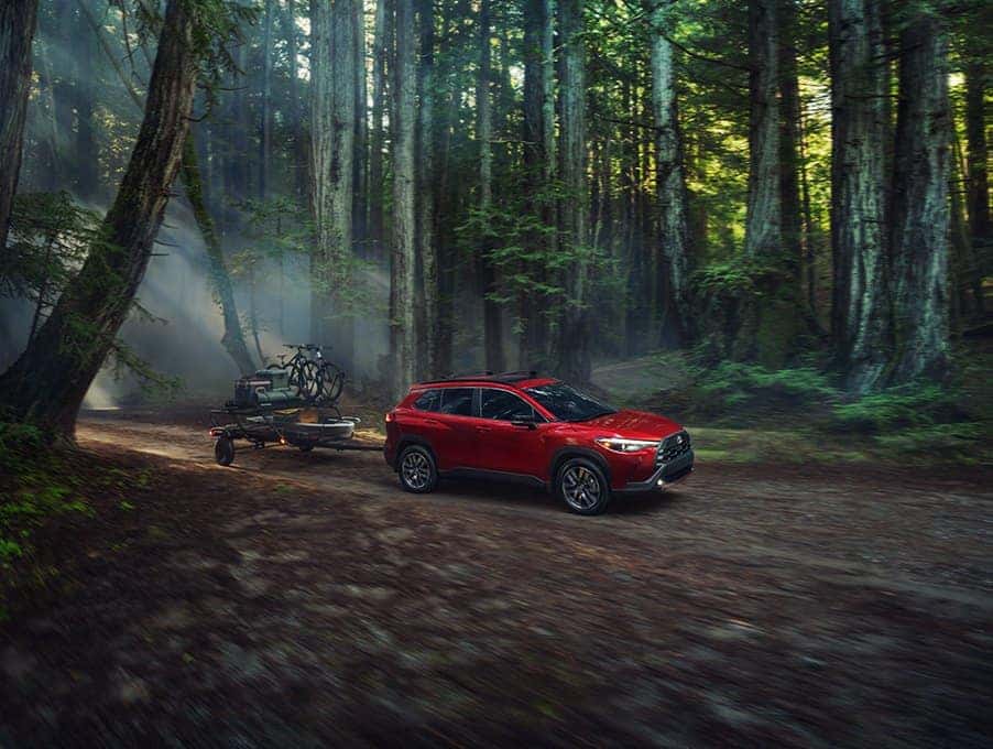 Red 2026 Toyota Corolla Cross XLE towing a small utility trailer on a forest road, available at Joseph Airport Toyota in Vandalia, Ohio