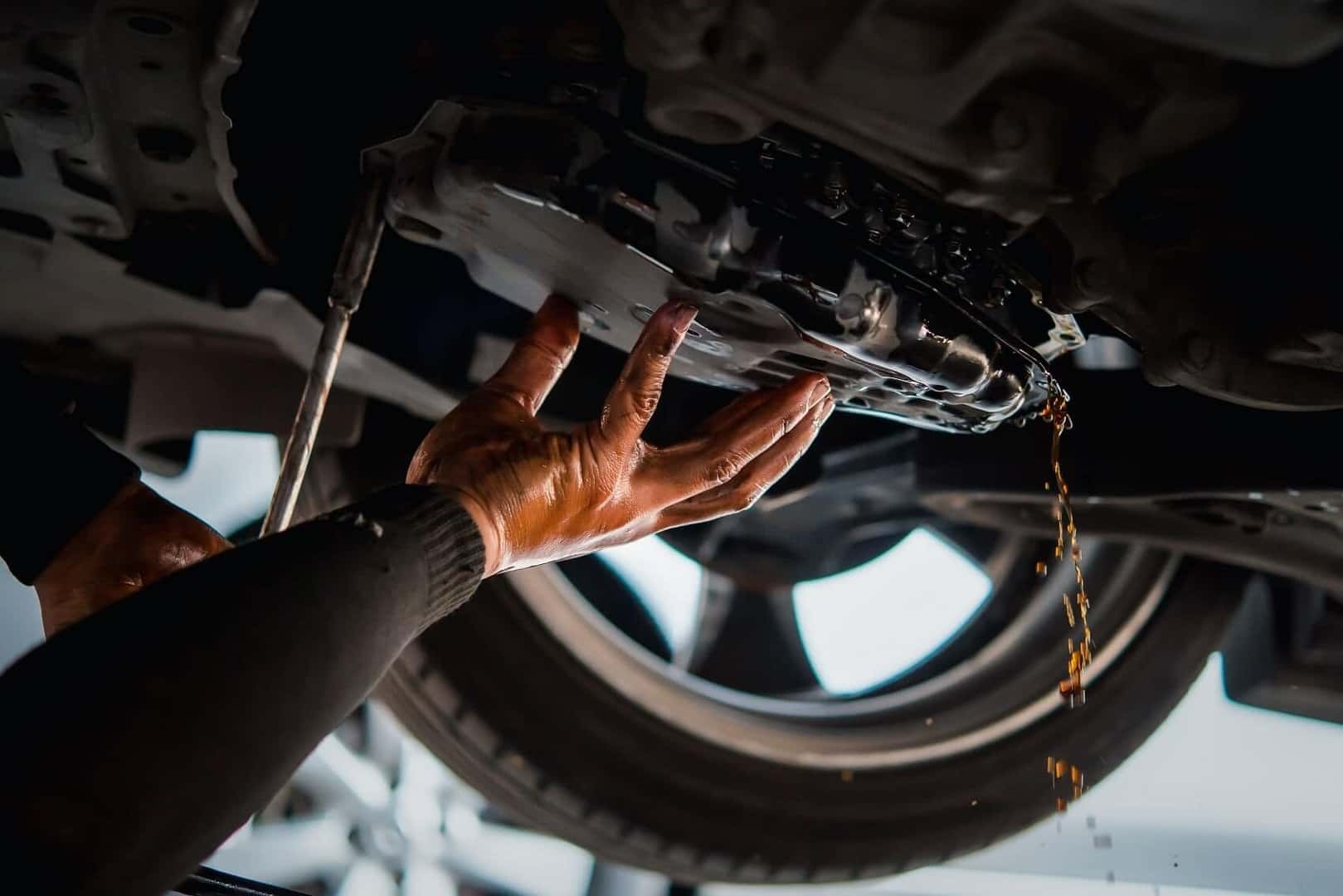 Undercarriage shot of a mechanic draining transmission fluid from a vehicle's transmission pan, amber fluid streaming down. Great service-department imagery.