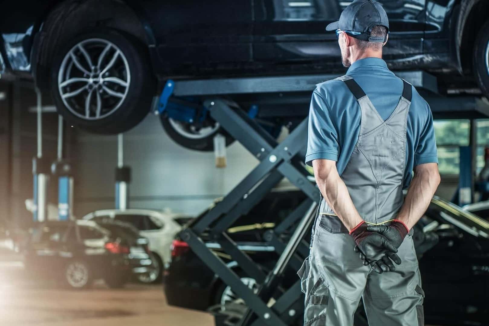 Technician in gray overalls and a cap standing with hands behind his back, looking up at a dark SUV raised on a scissor lift in a well-lit service bay