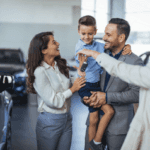 A happy family stands beside their new car at a dealership, smiling and celebrating their recent purchase together.