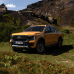 A bright orange pickup truck parked on a grassy field with rocky mountains and shrubs in the background under a partly cloudy sky.