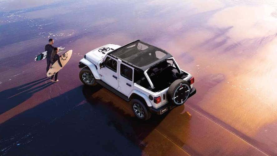 An overhead view of a white Jeep Wrangler on a beach with a couple with surfboards in front of it.