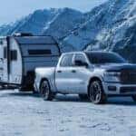 A silver RAM 1500 towing a trailer in a mountainous region covered in snow.