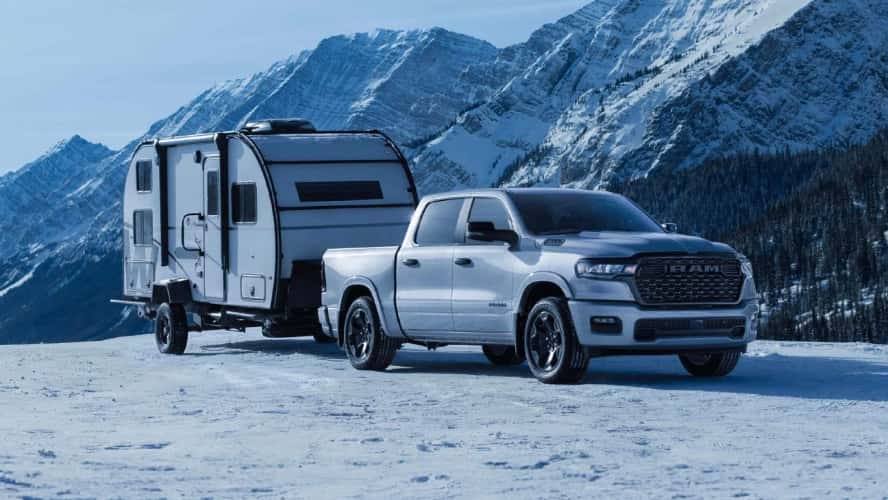 A silver RAM 1500 towing a trailer in a mountainous region covered in snow.