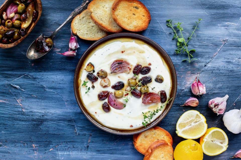 Flatlay of Greek feta cheese dip, Tirokafteri, with roasted olives, Italian garlic, red onions or shallots, drizzled with olive oil and garnished with lemon thyme
