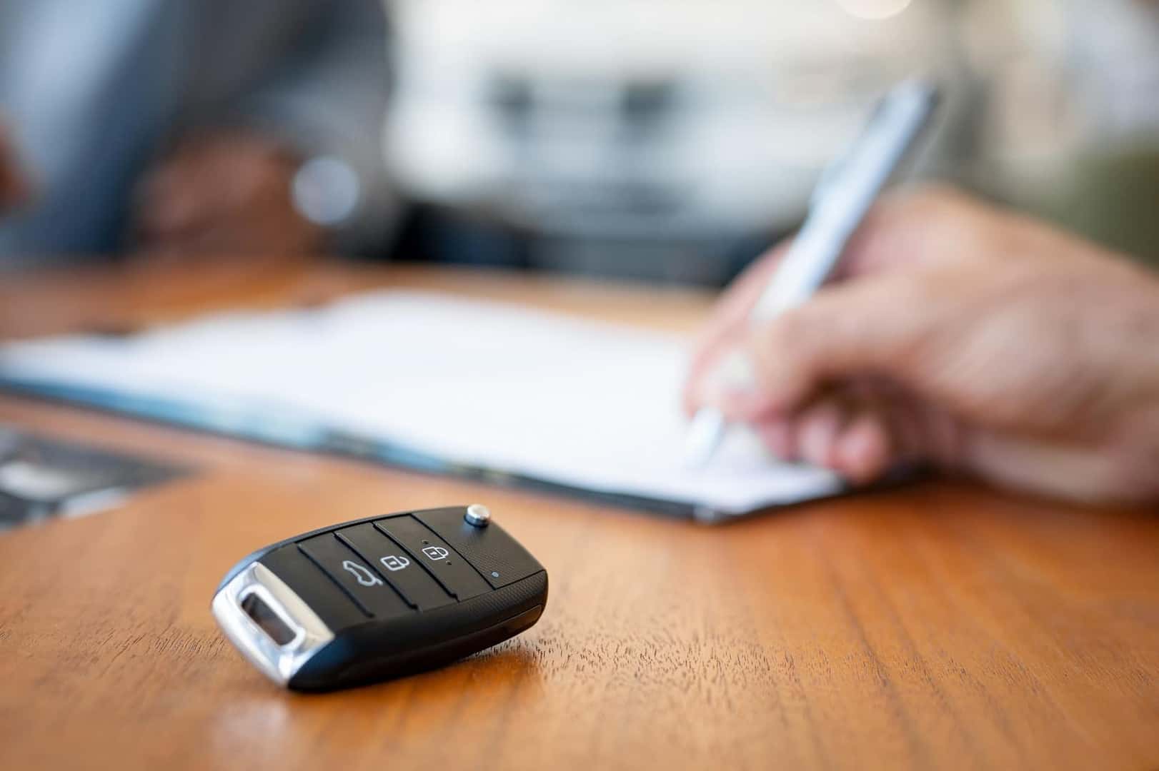Car,Keys,On,Desk,With,Man,Signing,Purchase,Documents,In