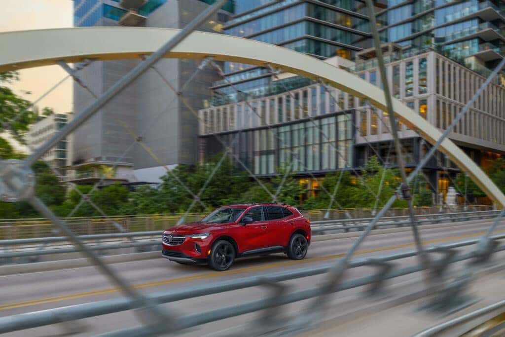Driver front 3/4 view of 2023 Buick Envision Essence in Cinnabar Metallic driving on bridge. Due to current supply-chain shortages, certain features shown have limited or late availability, or are no longer available. See the window label or a dealer regarding the features on an individual vehicle.