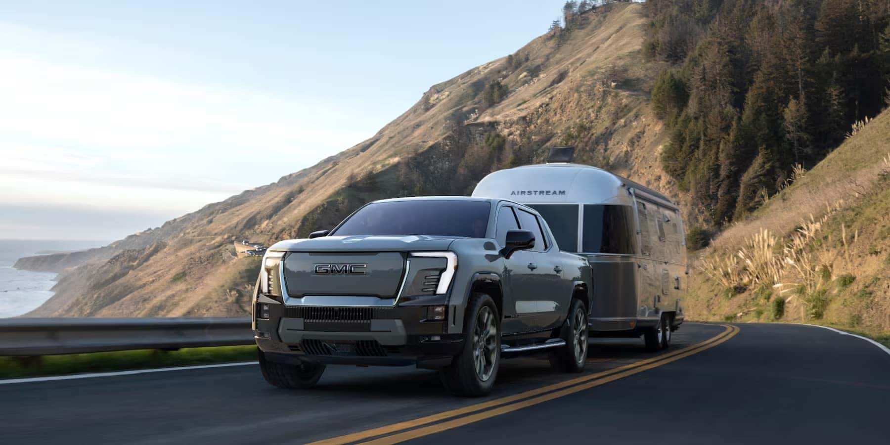 2025 GMC Sierra EV Denali first Edition towing a camper up a coastal mountain road.
