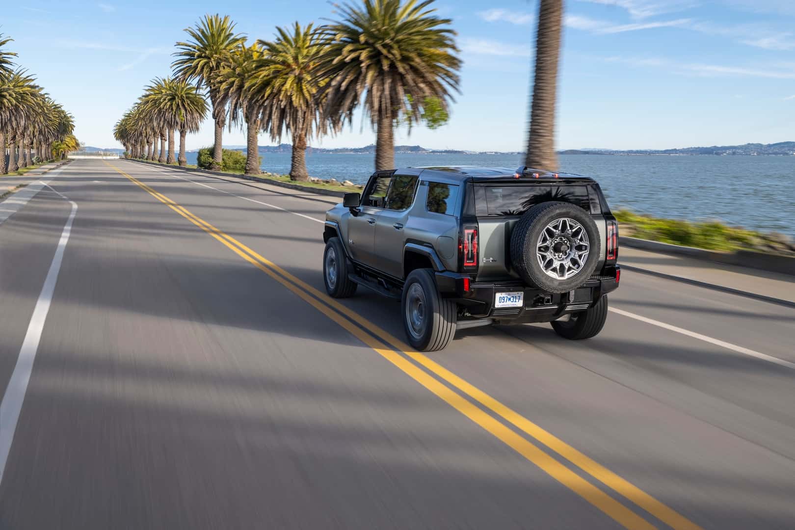2024 GMC HUMMER EV SUV drives down a coastal road with palm trees.