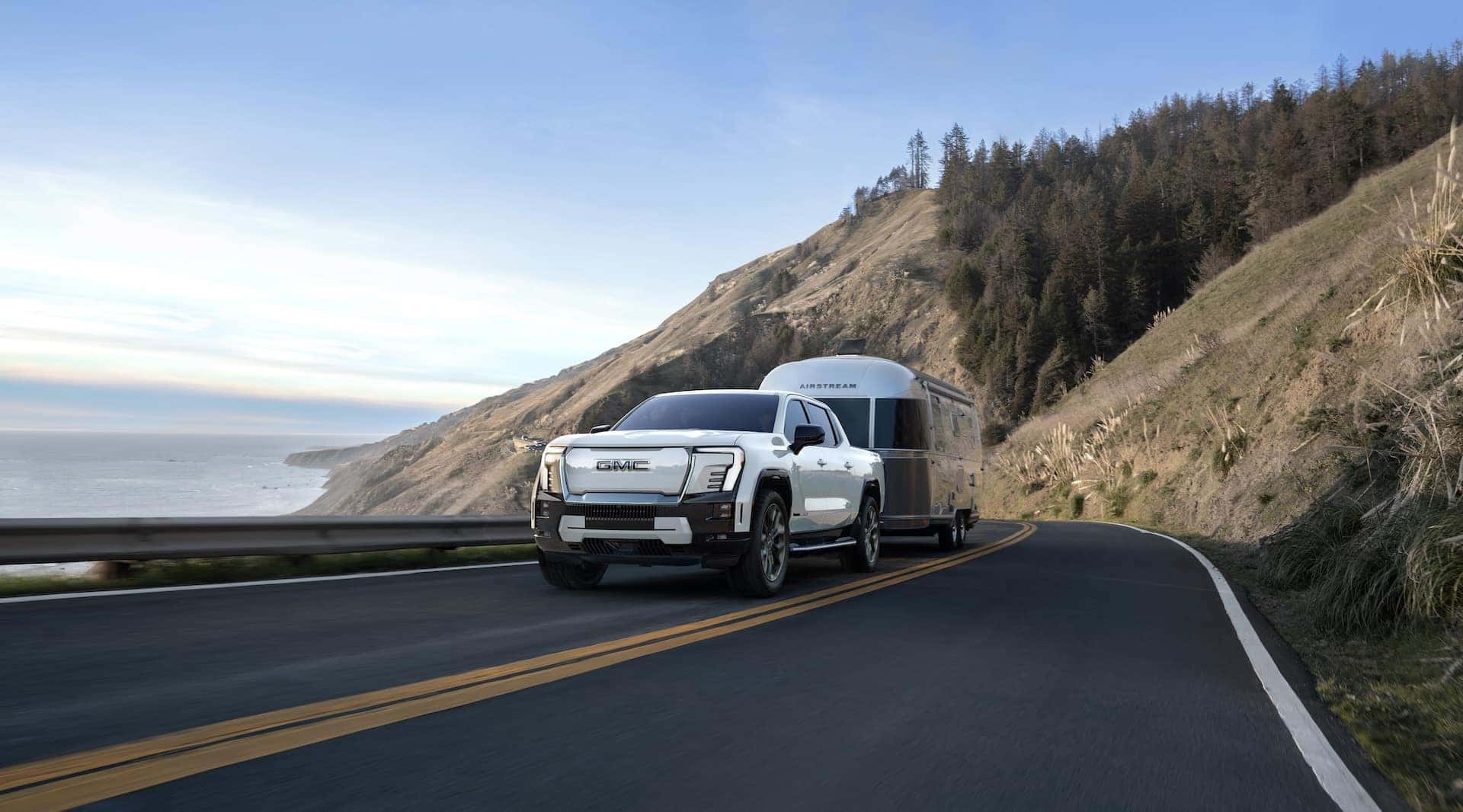 Front 3/4 view of the 2025 GMC Sierra EV Denali towing a trailer on a seaside road.