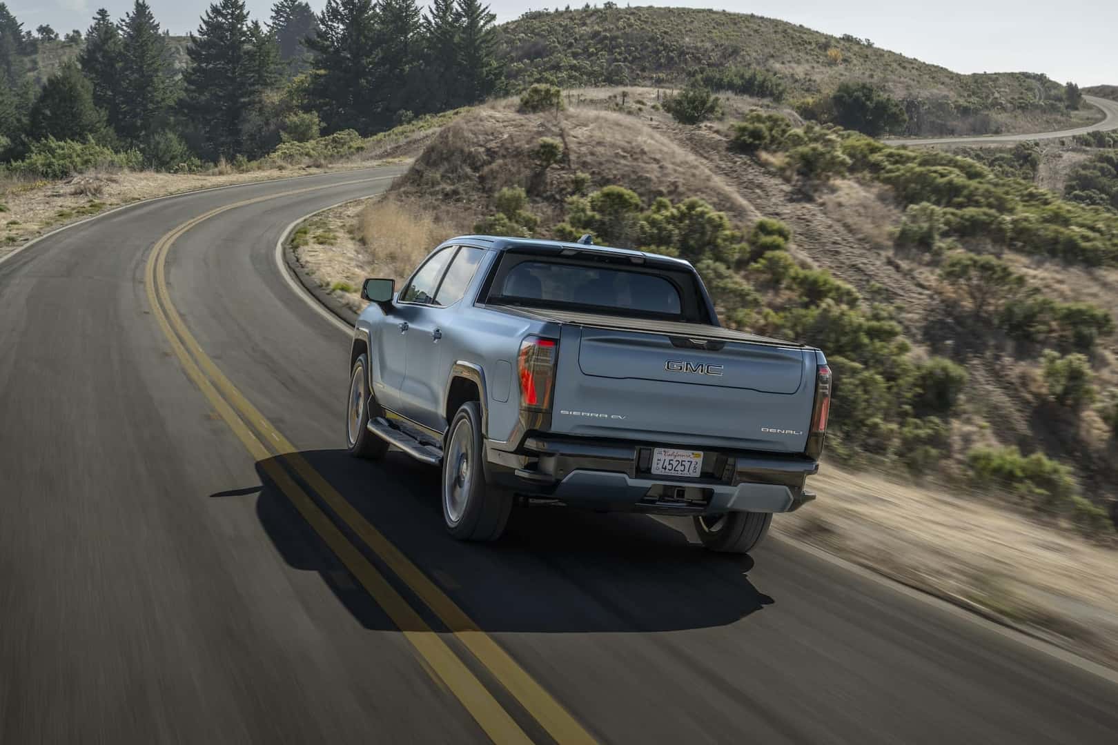 Rear shot of 2025 GMC Sierra EV Denali in Moonlight Matte driving uphill on two lane divided road.