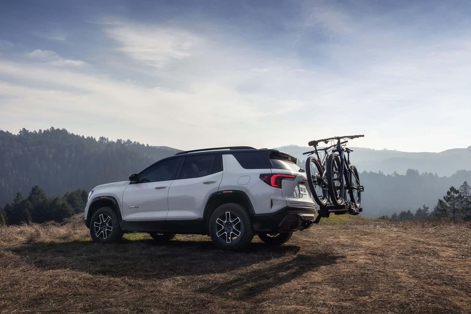 The Comfortable And Competent 2026 GMC Terrain AT4 With Bikes On Its Back, Cruising Through The Grass Hills Near Vast Mountains And Forests As Far As The Eye Can See