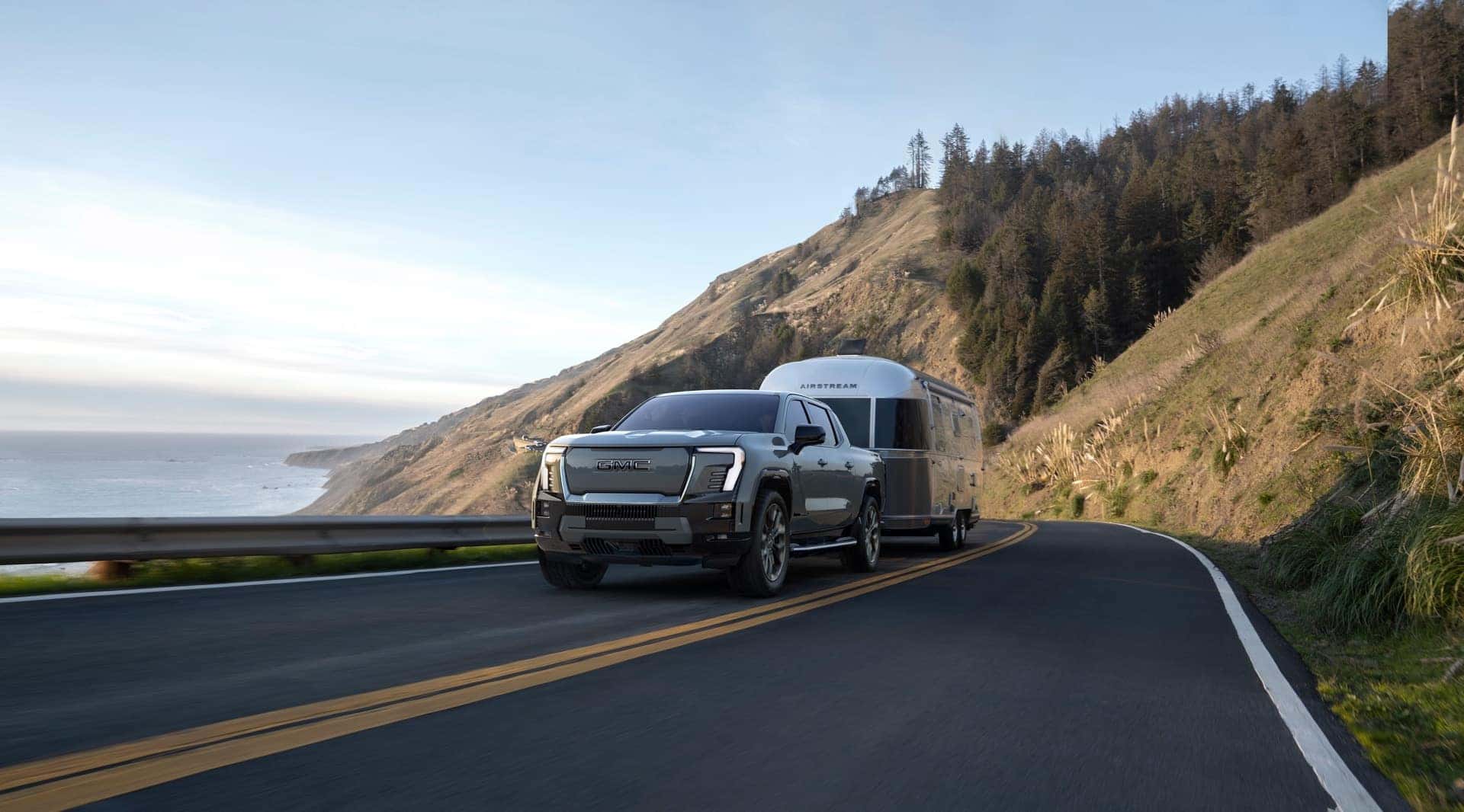 The Vigorous And Powerful 2024 GMC Sierra EV Denali Edition Towing A Trailer With Ease, In View Of A Blue Sky And Vast Mountains As The Car Travels On A Road