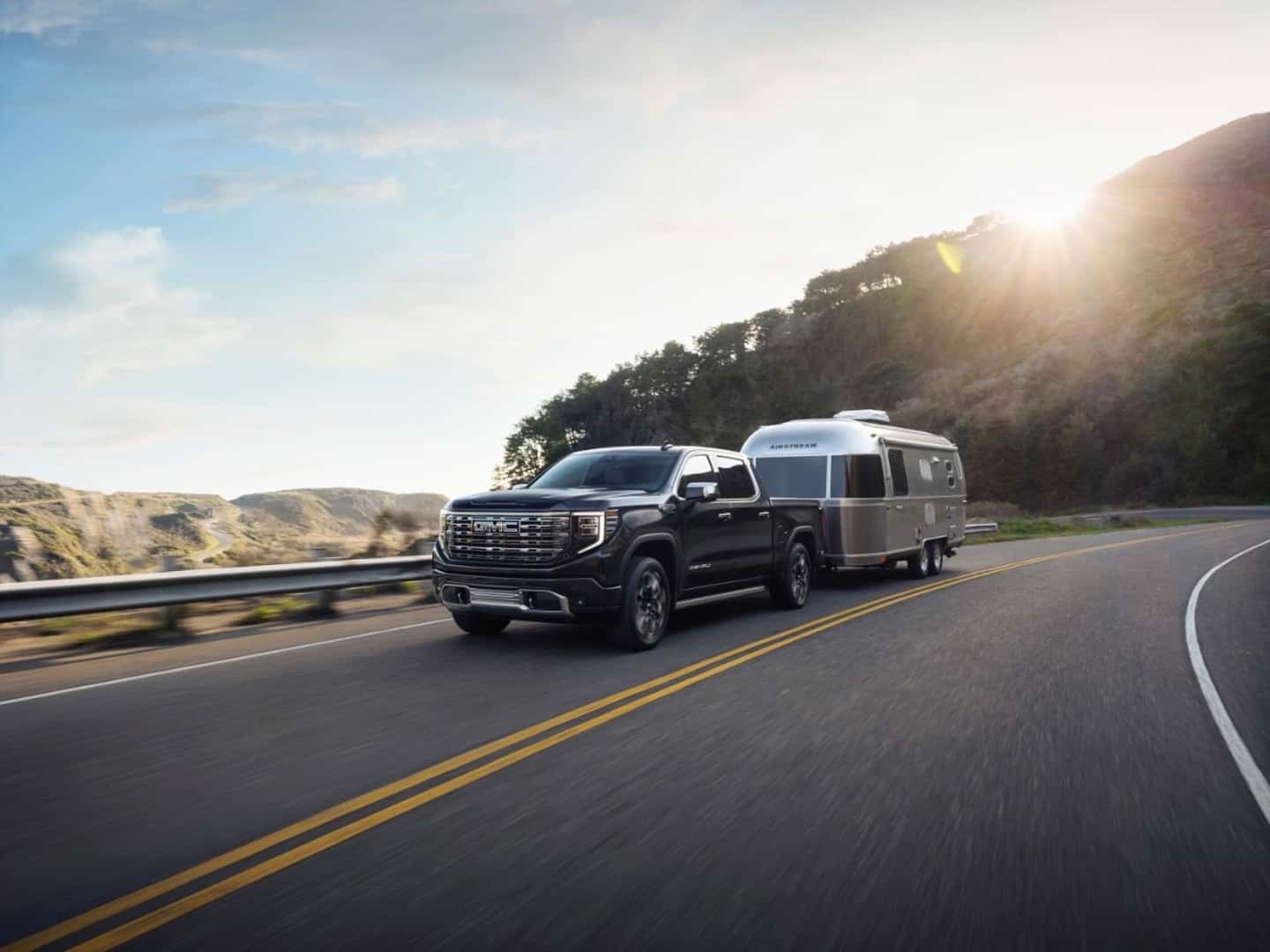 The Muscular And Sturdy 2026 GMC Sierra Denali Ultimate 1500 Towing A Large And Metallic Trailer, With The Sun Shining Down From A Bright Blue Sky With A Few Sparse Clouds
