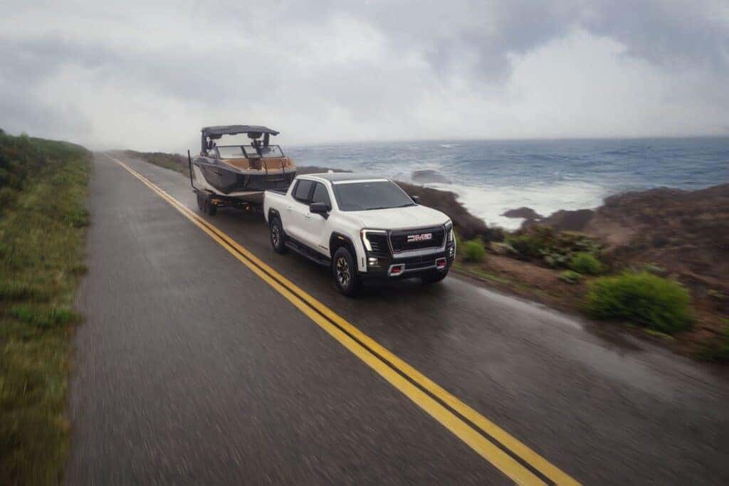 The Strong And Robust 2026 GMC Sierra EV AT4 Towing A Boat Trailer With Ease, Under A Clouded Sky On Wet Roads