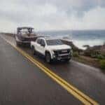 The Strong And Robust 2026 GMC Sierra EV AT4 Towing A Boat Trailer With Ease, Under A Clouded Sky On Wet Roads