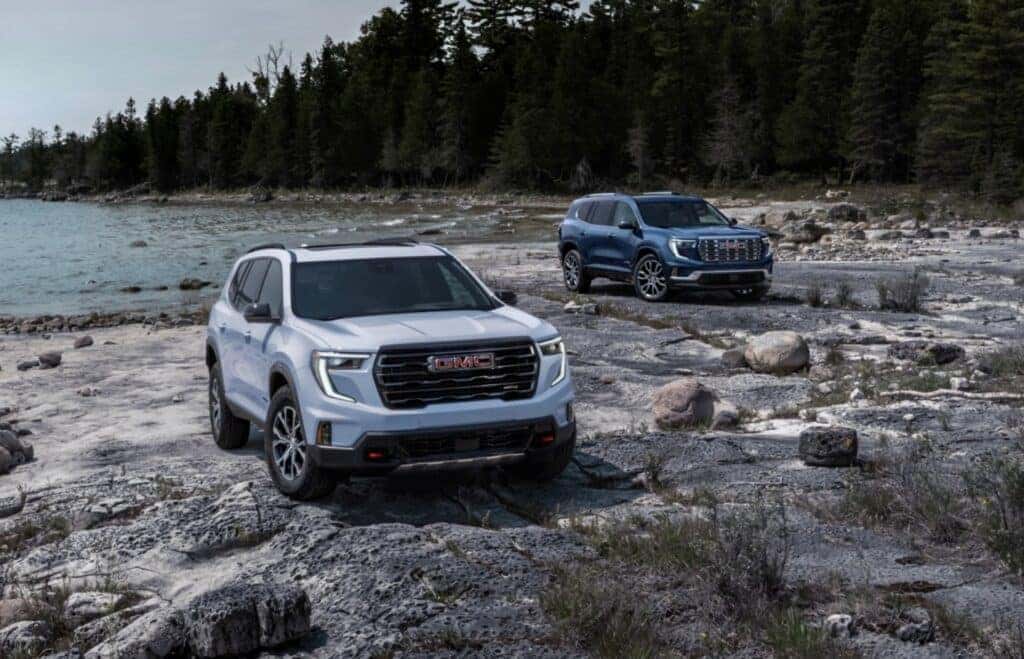 Two Adventurous 2025 GMC Acadia AT4s Parked On A Rocky Beach, Near Trees