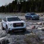 Two Adventurous 2025 GMC Acadia AT4s Parked On A Rocky Beach, Near Trees