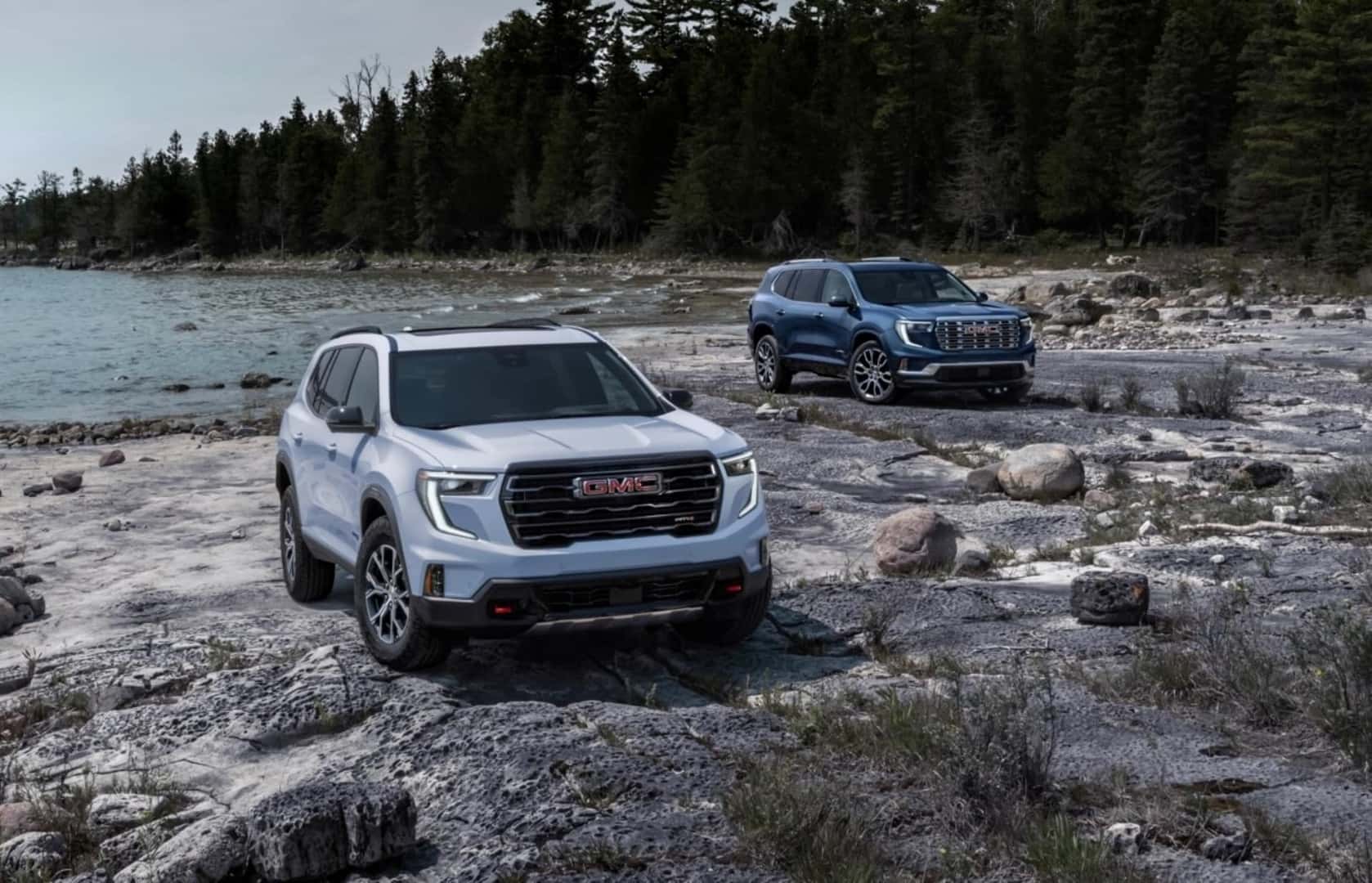 Two Adventurous 2025 GMC Acadia AT4s Parked On A Rocky Beach, Near Trees