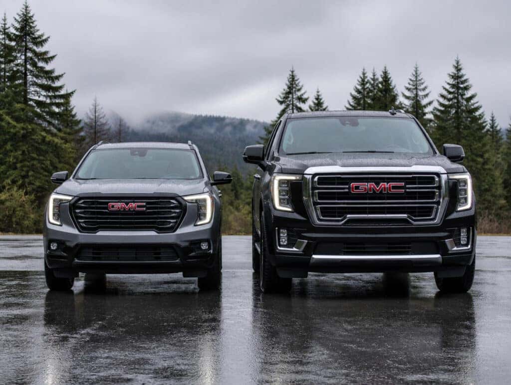 2026 GMC Acadia and 2026 GMC Yukon parked side by side on wet pavement with Pacific Northwest evergreens and gray sky