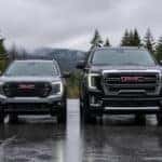 2026 GMC Acadia and 2026 GMC Yukon parked side by side on wet pavement with Pacific Northwest evergreens and gray sky