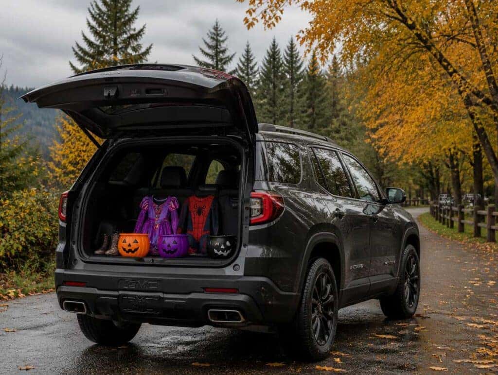 2026 GMC Acadia with liftgate open at a fall festival, Halloween costumes and candy buckets in the cargo area, evergreen and maple trees in a wet Pacific Northwest park setting